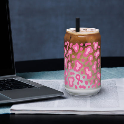 A clear glass can-shaped tumbler with a pink leopard print pattern, filled with an iced coffee drink and topped with a black straw, set on a newspaper next to a laptop.