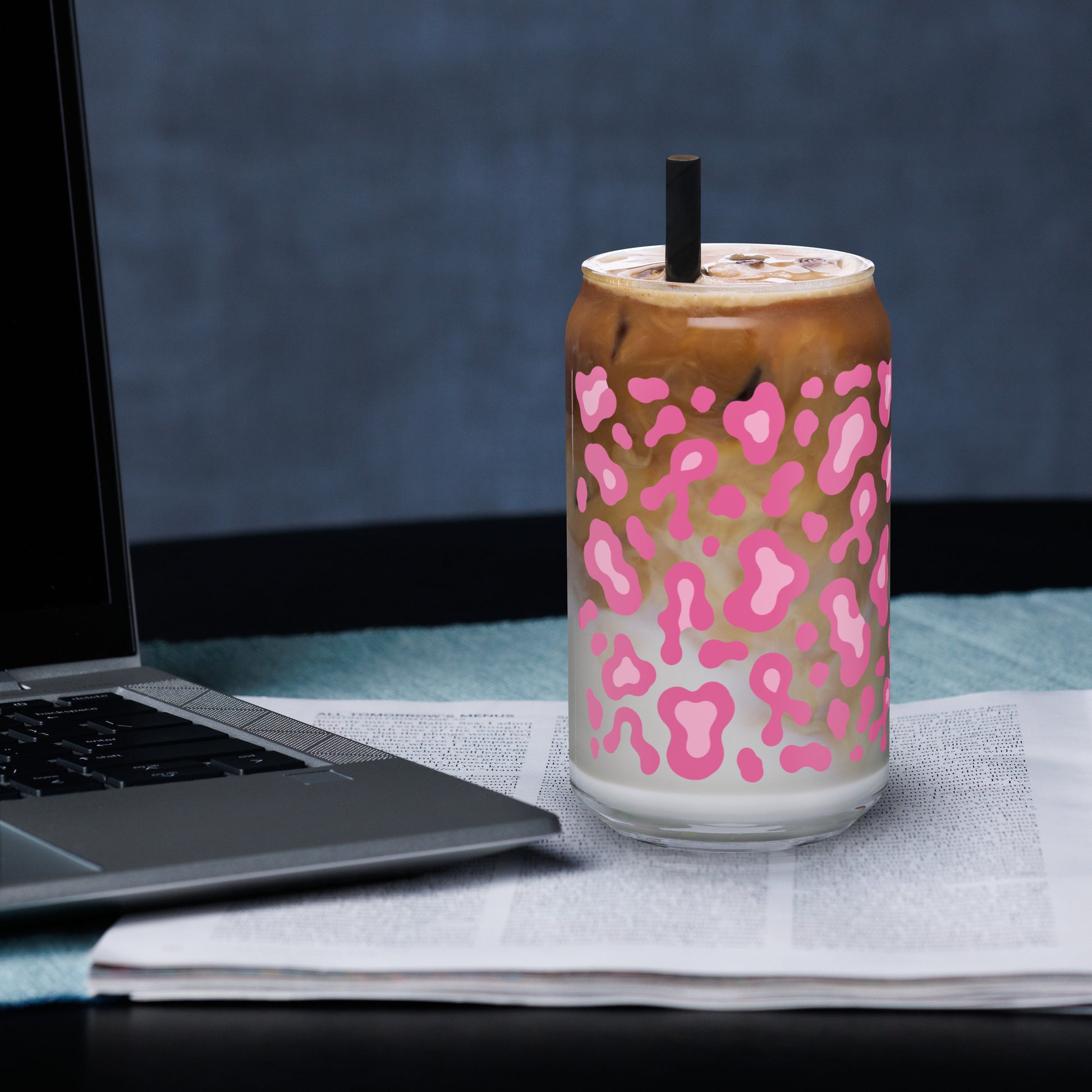 A clear glass can-shaped tumbler with a pink leopard print pattern, filled with an iced coffee drink and topped with a black straw, set on a desk next to an open laptop and newspaper.