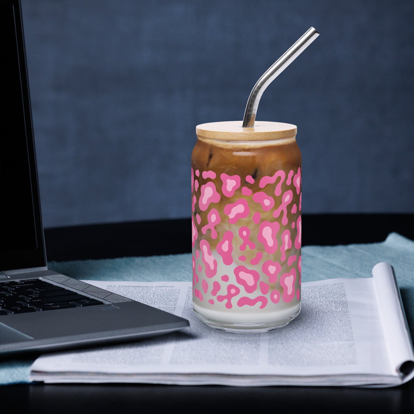 A clear glass can-shaped tumbler with a pink leopard print pattern, filled with an iced coffee drink and topped with a light wood lid and a bent metal straw. The tumbler sits on a newspaper next to a laptop against a dark blue-gray background.