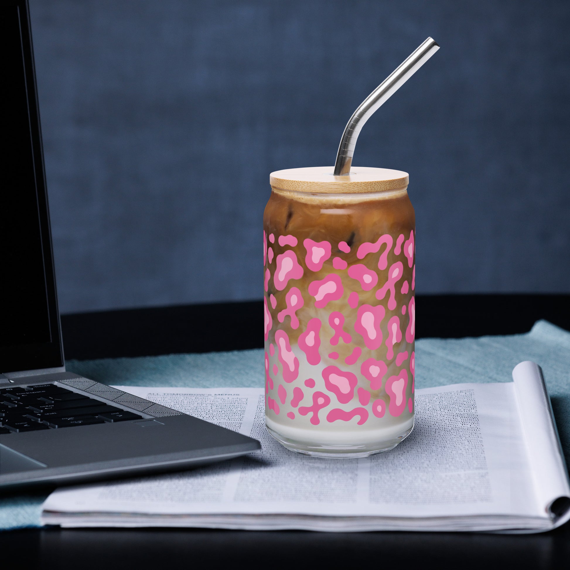 A clear glass can-shaped tumbler with a pink leopard print pattern, filled with an iced coffee drink and topped with a light wood lid and a bent metal straw. The tumbler sits on a newspaper next to a laptop against a dark blue-gray background.