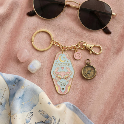 A pastel pink and gold Cancer zodiac keychain charm with a compass and pink gemstone charms, styled on a pink velvet fabric with sunglasses, crystals, and a blue-and-white starry scarf.