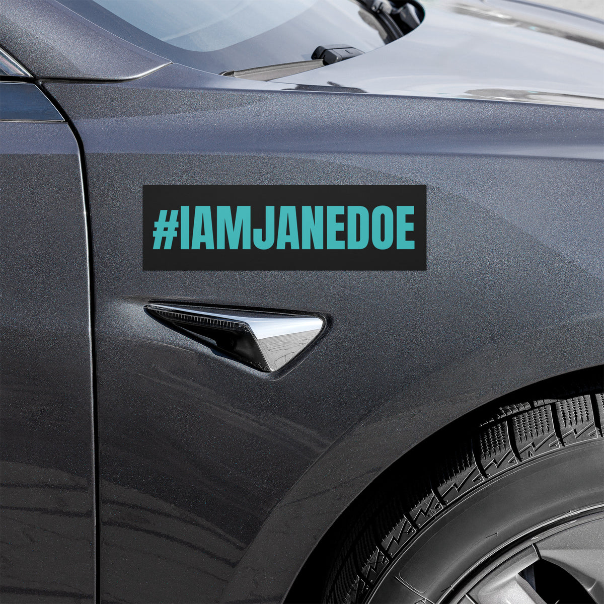 A rectangular black car decal with the text '#IAMJANEDOE' in bright teal, shown applied to the side door of a dark gray vehicle.