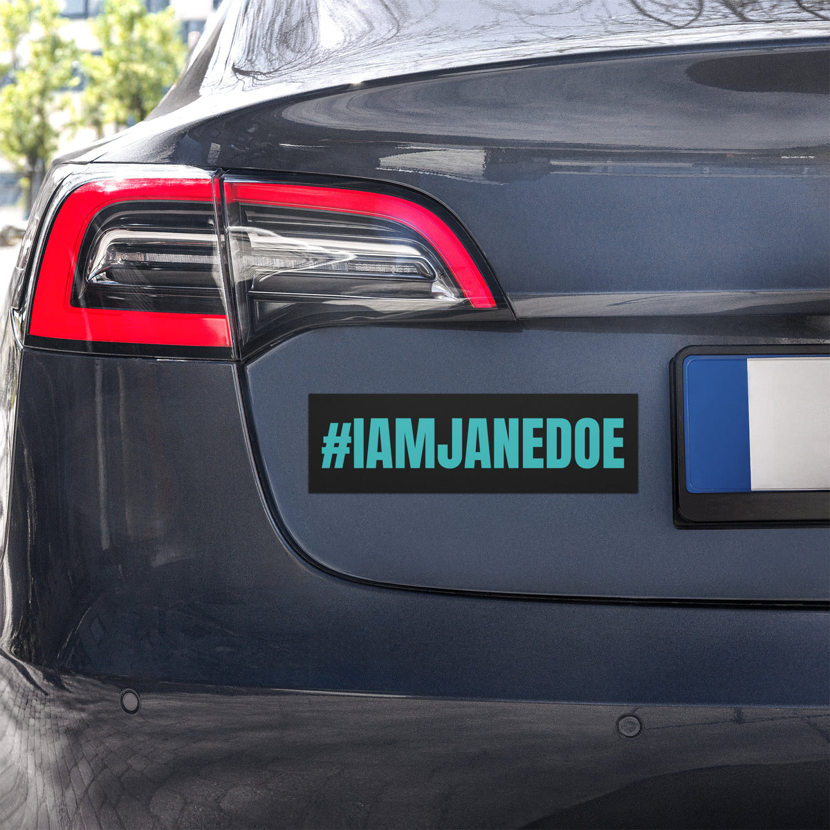 A rectangular black bumper sticker with the text '#IAMJANEDOE' in bright teal, shown applied to the rear bumper of a dark gray vehicle. A blue and white license plate is visible on the right.