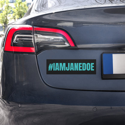 A rectangular black bumper sticker with the text '#IAMJANEDOE' in bright teal, shown applied to the rear bumper of a dark gray vehicle. A blue and white license plate is visible on the right.