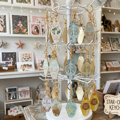A white multi-tier display stand holding multiple decorative star-shaped keychains with gold-tone rings and colorful printed designs, set on a rustic wooden table inside a shop. A sign reading “STAR-CROSSED KEYCHAINS” is visible nearby.