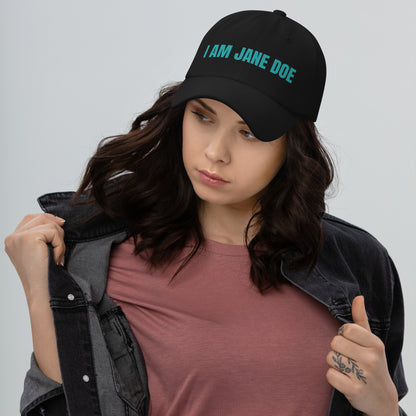 Woman wearing a black cap with 'I AM JANE DOE' embroidered in teal thread on a gray background