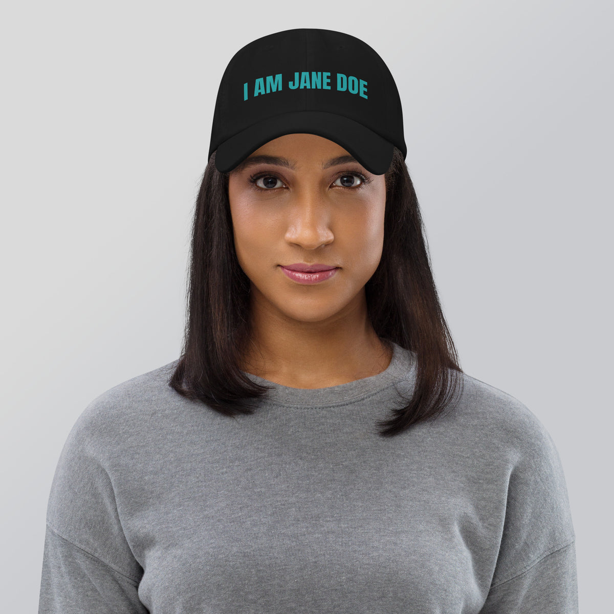 Woman wearing a black cap with a 'I AM JANE DOE' embroidered in teal thread, on a gray background