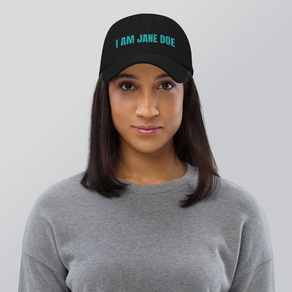 Woman wearing a black cap with a 'I AM JANE DOE' embroidered in teal thread, on a gray background