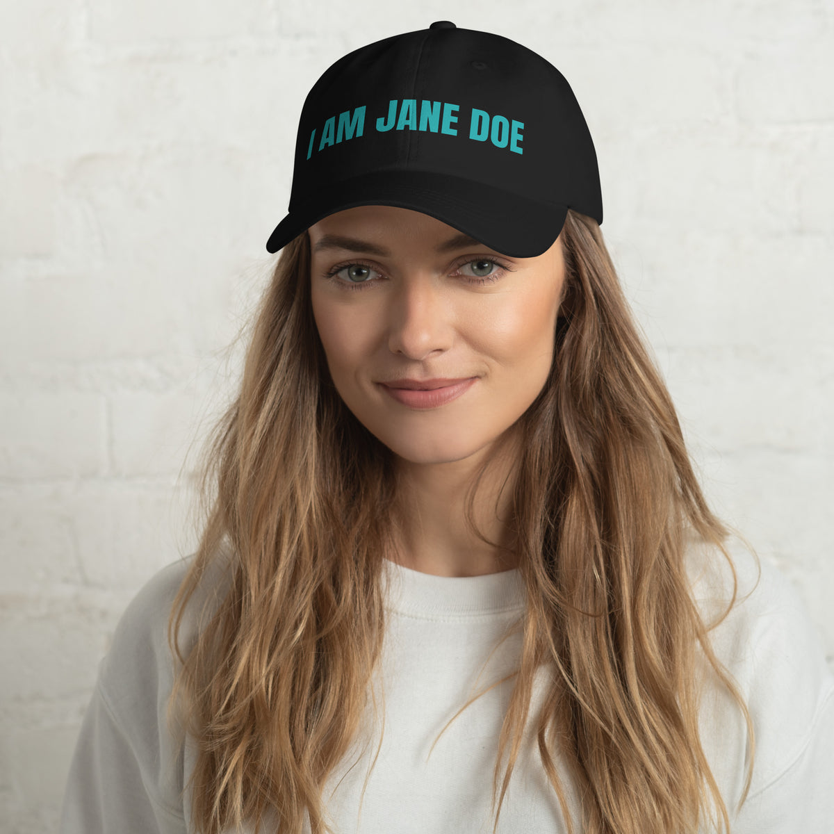 Person wearing a black cap with 'I AM JANE DOE' text embroidered in teal thread on a white background