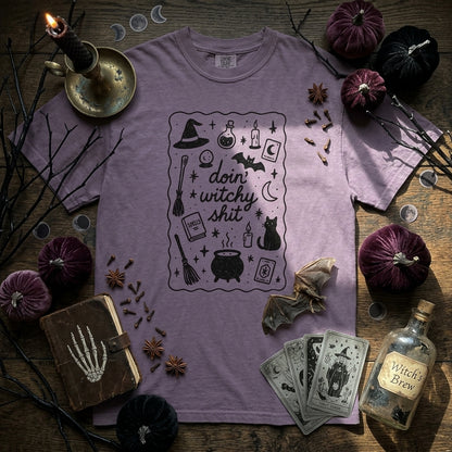 A purple short-sleeve t-shirt with a black graphic print reading 'doin' witchy shit' surrounded by Halloween-themed props, styled in a flat-lay on a rustic wooden background.