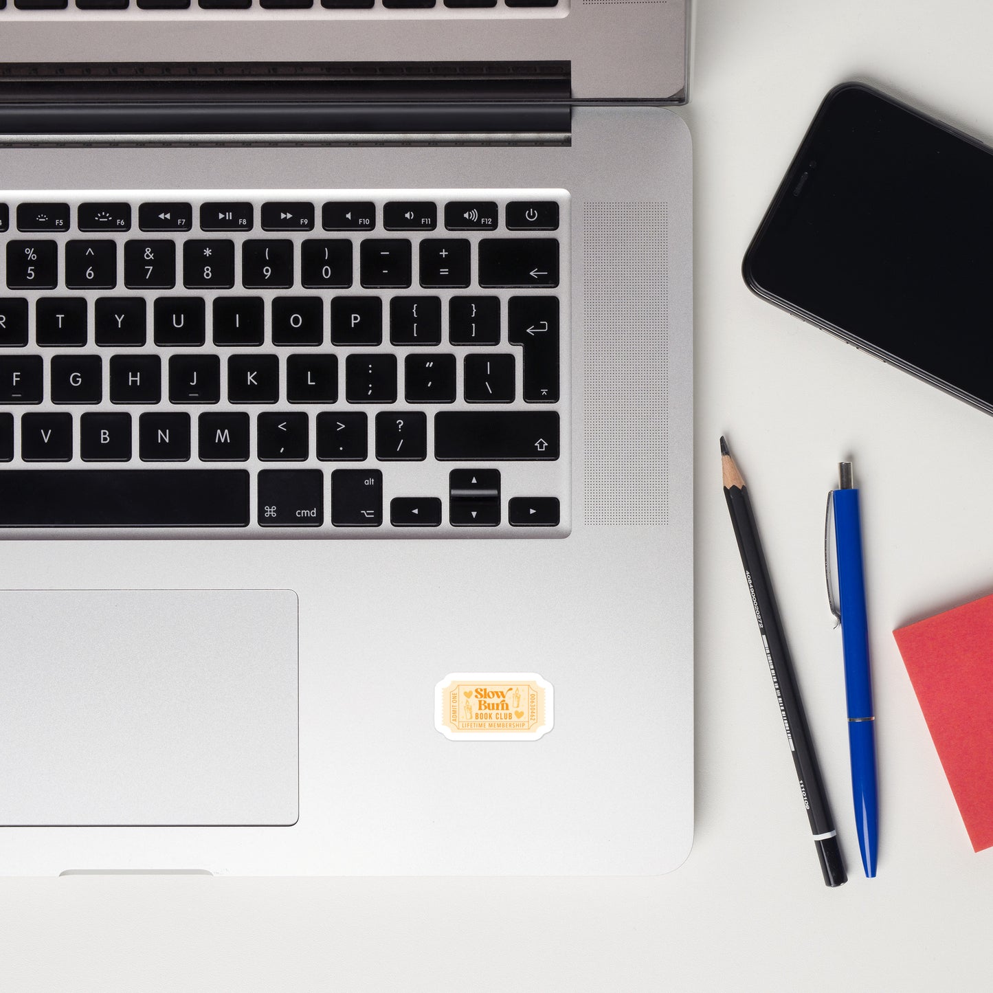 Laptop with a yellow book trope lifetime member ticket stub sticker on a desk with a notebook, mouse, pencil, and plants.