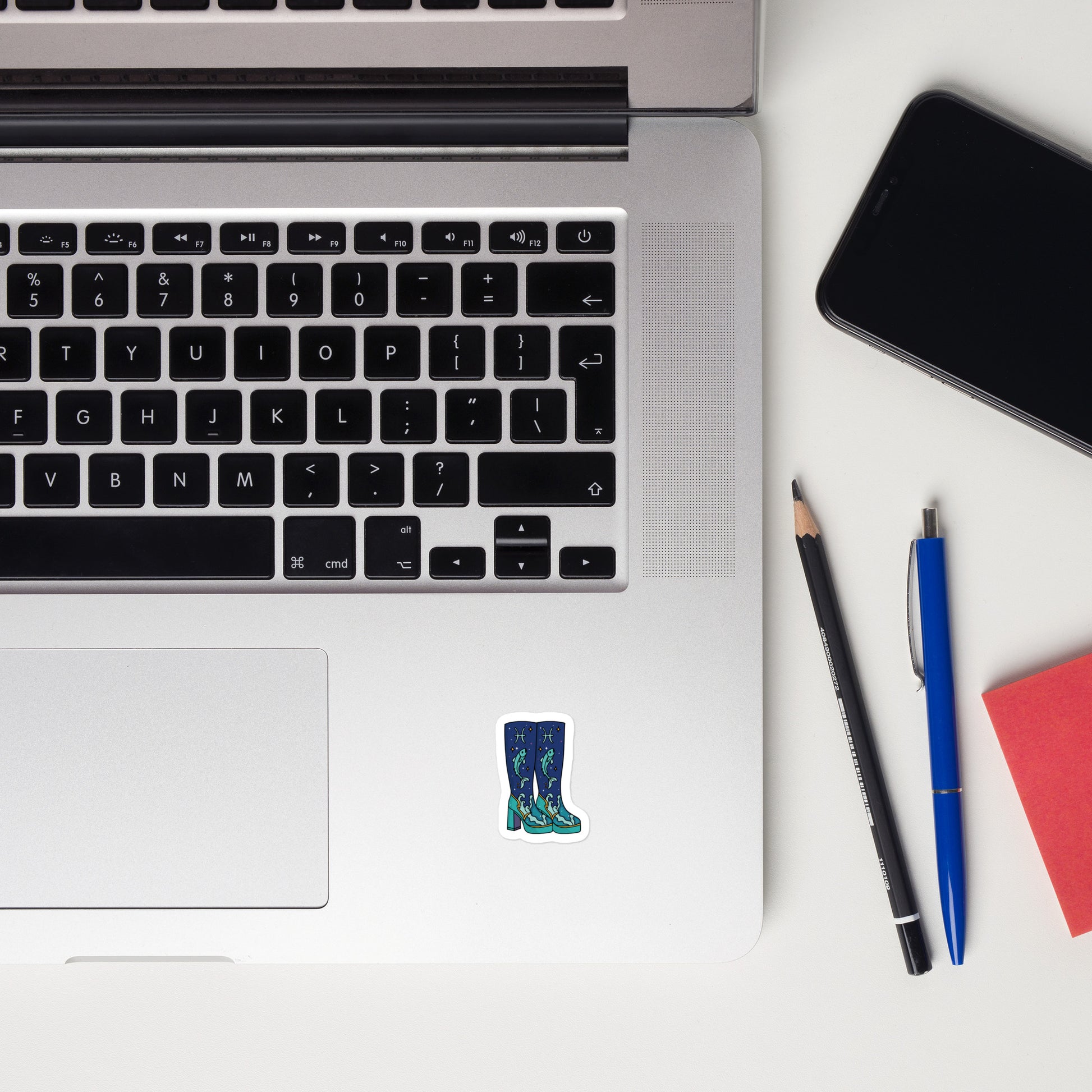A close-up flat-lay of a silver laptop keyboard with a small die-cut sticker of blue rain boots on the palm rest, surrounded by a black smartphone, a pencil, a blue pen, and a red notebook on a white surface.