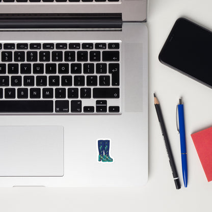 A close-up flat-lay of a silver laptop keyboard with a small die-cut sticker of blue rain boots on the palm rest, surrounded by a black smartphone, a pencil, a blue pen, and a red notebook on a white surface.