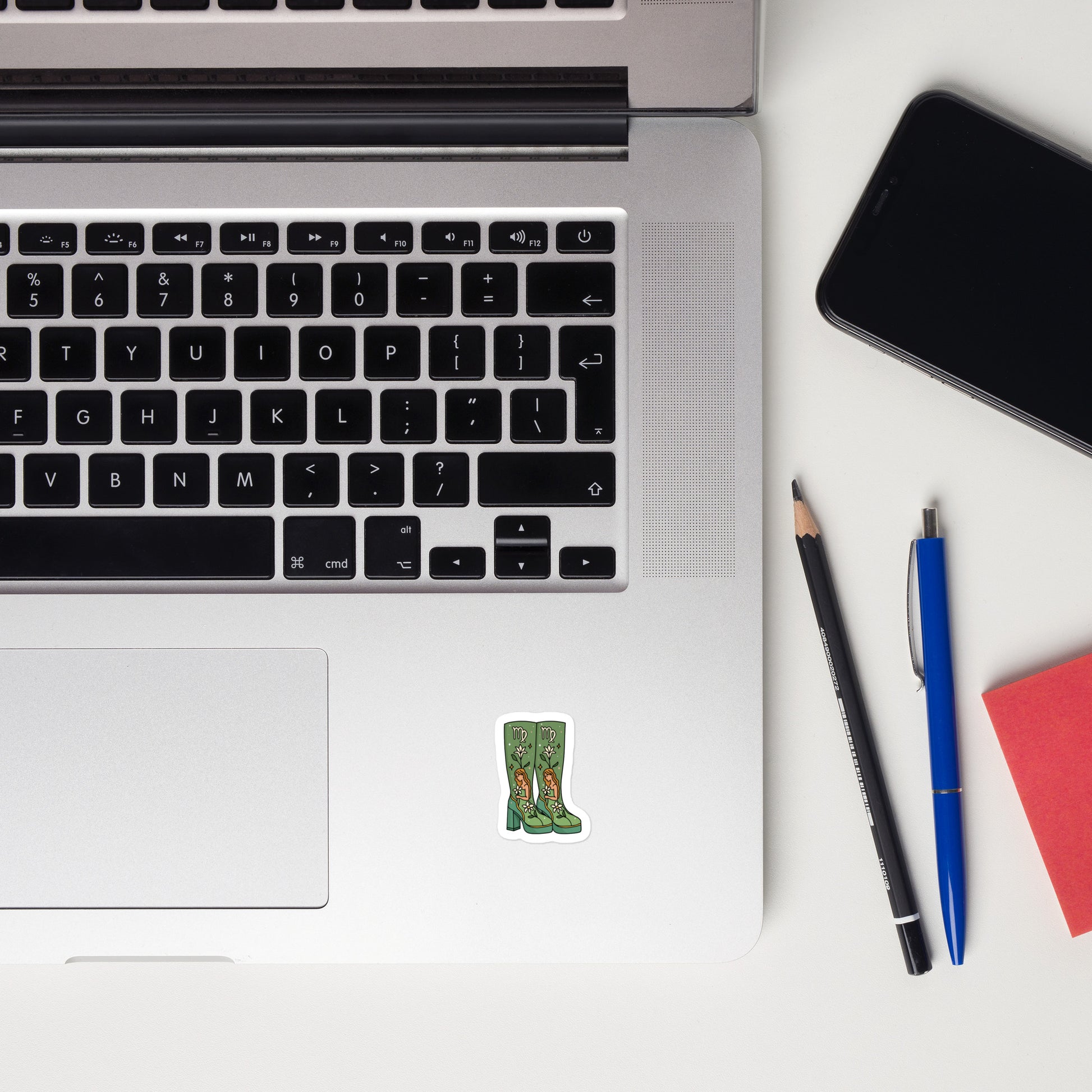 Laptop with a sticker of platform boots with a zodiac inspired design, smartphone, and writing utensils on a white surface