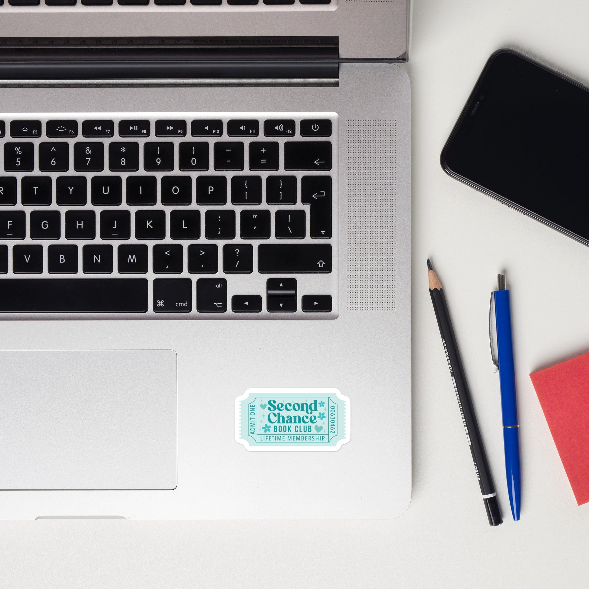 Laptop with a teal book trope lifetime member ticket stub sticker on a desk with a notebook, mouse, pencil, and plants.