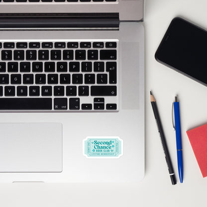 Laptop with a teal book trope lifetime member ticket stub sticker on a desk with a notebook, mouse, pencil, and plants.