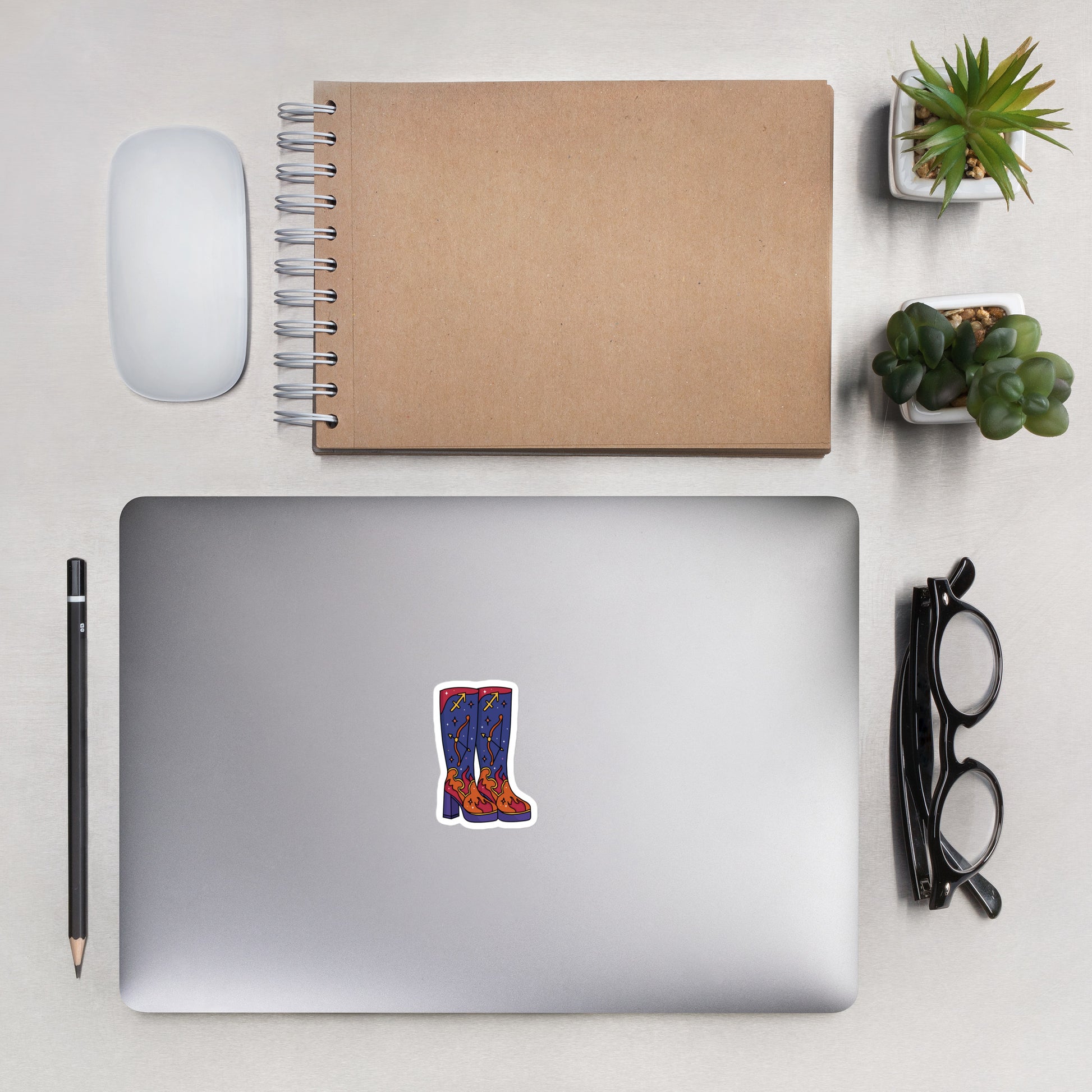 A styled flat-lay workspace scene featuring a silver laptop with a die-cut sticker of blue jeans and red boots on the lid, alongside a brown kraft spiral notebook, a white mouse, a black pencil, black eyeglasses, and two small potted succulents on a light gray surface.
