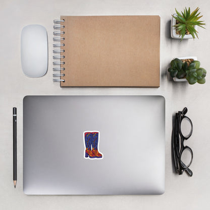A styled flat-lay workspace scene featuring a silver laptop with a die-cut sticker of blue jeans and red boots on the lid, alongside a brown kraft spiral notebook, a white mouse, a black pencil, black eyeglasses, and two small potted succulents on a light gray surface.