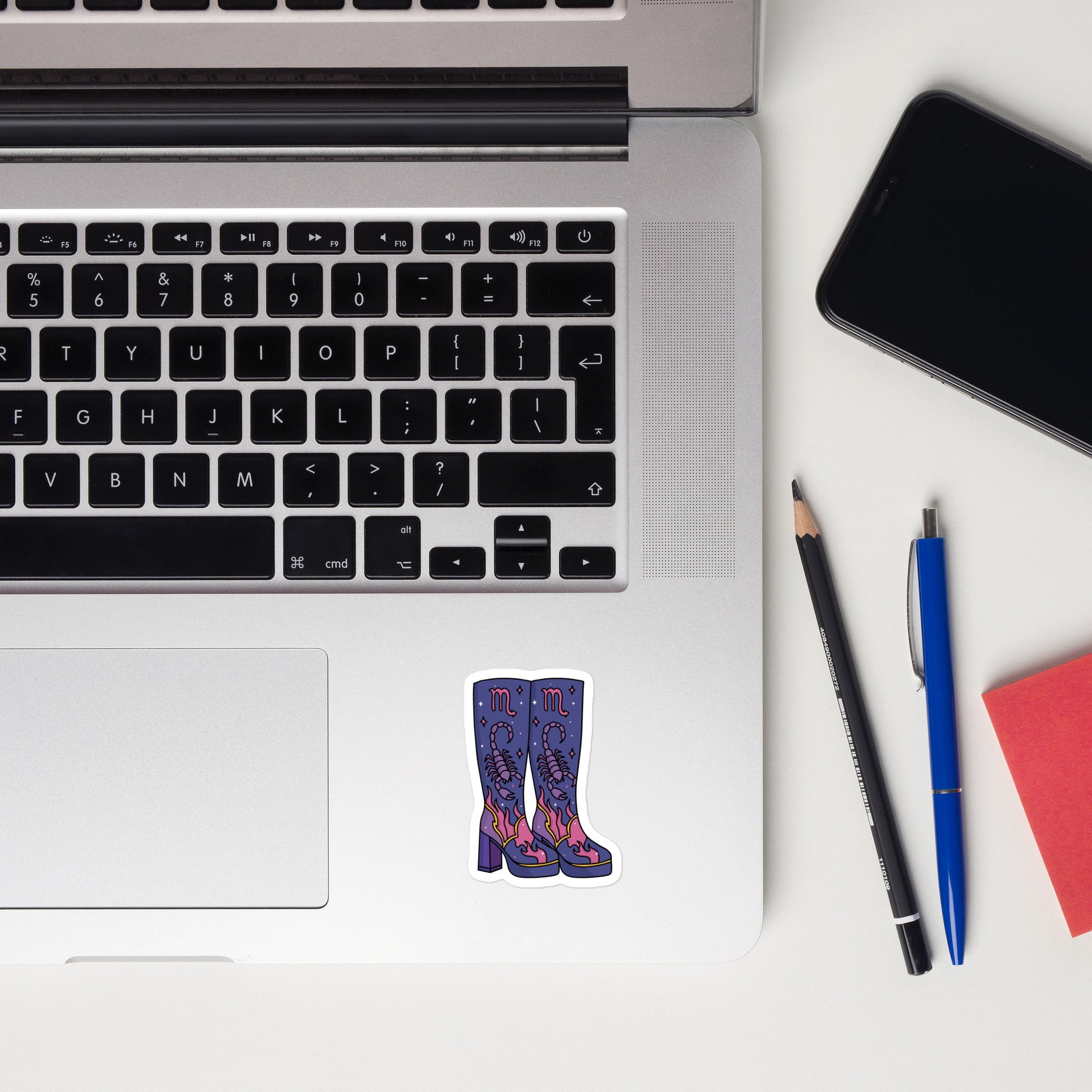 A flat-lay view of a silver laptop on a white desk, featuring a die-cut sticker of purple patterned high-heeled boots on the bottom left of the keyboard deck. Surrounding the laptop are a black smartphone, a blue pen, a black pencil, and a red notebook.