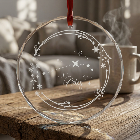 A clear faceted glass Christmas ornament hanging from a red ribbon, engraved with an Aries zodiac constellation design, resting on a rustic wooden table with a blurred living room background and a mug.