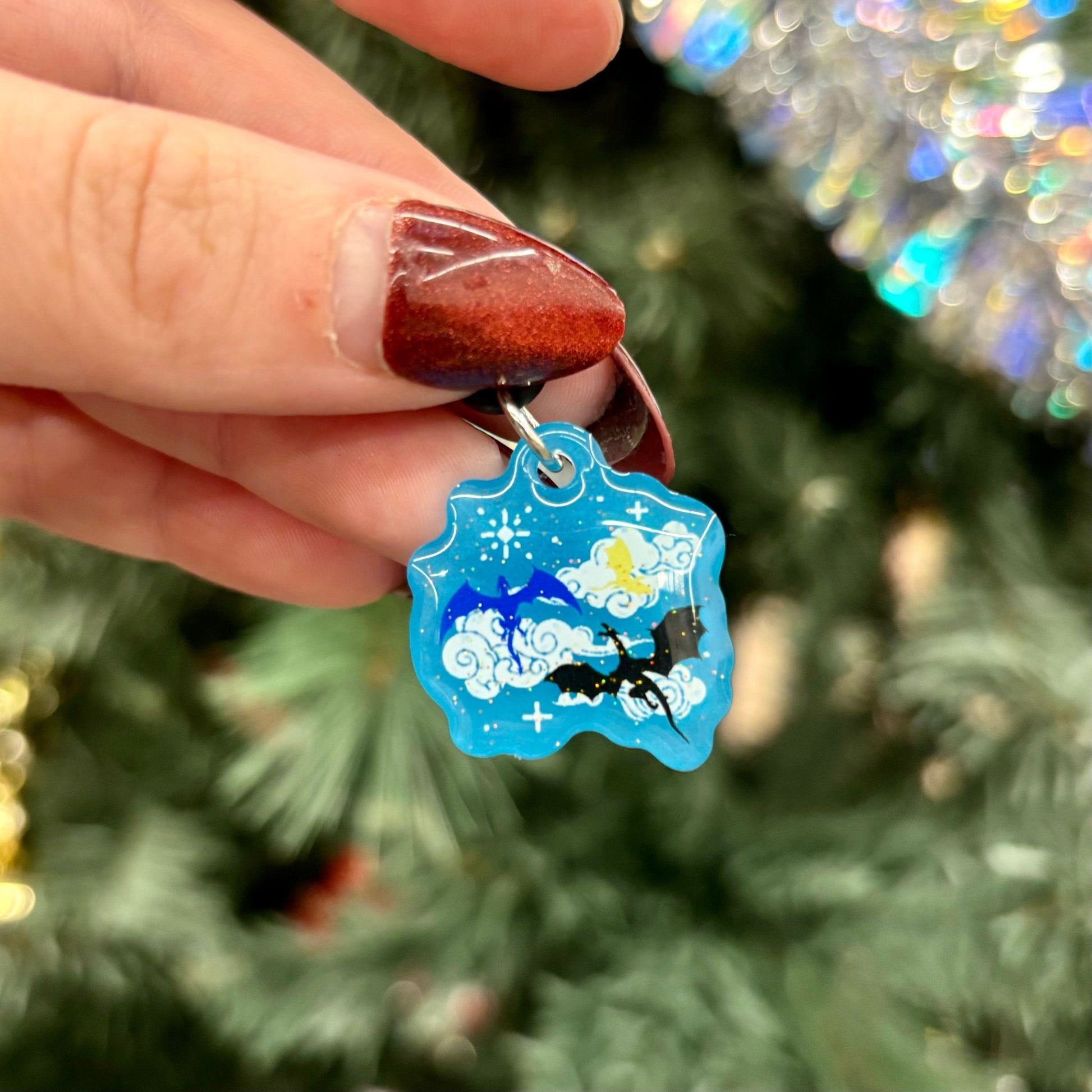 Hand holding a blue decorative Kindle Charm featuring 3 dragons, one blue, one gold, and one black, and a starry sky background against a blurred Christmas tree background.