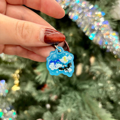 Hand holding a blue kindle charm featuring 3 dragons, one blue, one gold, and one black with a star design against a festive background.
