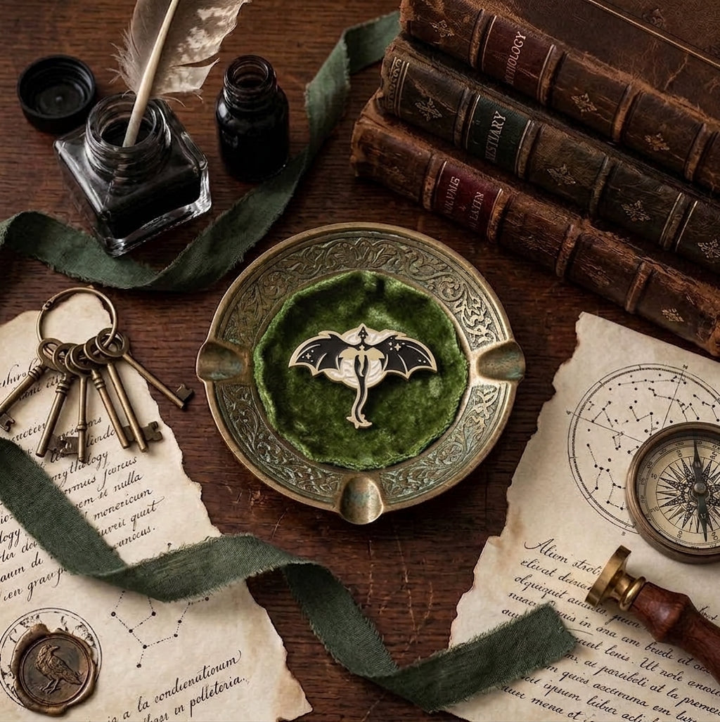 A decorative metal bronze trinket tray with an enamel dragon pin resting in it, displayed on a dark wooden table surrounded by vintage props including old books, keys, parchment, ink, and a compass