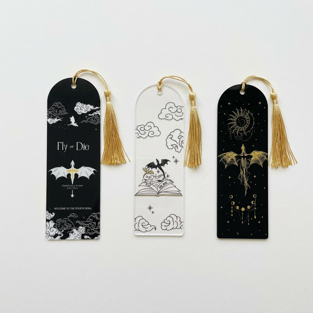 Three decorative acrylic bookmarks with gold tassels are displayed on a clean white background. The left bookmark is black with white cloud and dragon illustrations and the text 'Fly or Die', the middle, clear with dragons flying from an open book with the text 'FLY OR DIE', and the right, black with a gold dragon and celestial design