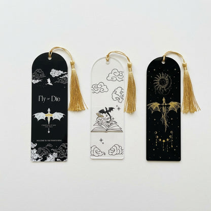 Three decorative acrylic bookmarks with gold tassels are displayed on a clean white background. The left bookmark is black with white cloud and dragon illustrations and the text 'Fly or Die', the middle, clear with dragons flying from an open book with the text 'FLY OR DIE', and the right, black with a gold dragon and celestial design