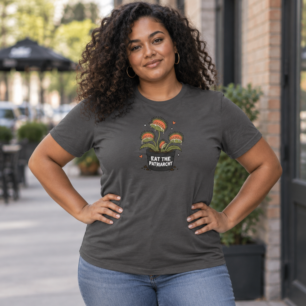 A woman with curly hair stands outdoors wearing a dark charcoal gray graphic t-shirt with a colorful venus fly trap floral design and the text 'EAT THE PATRIARCHY'.