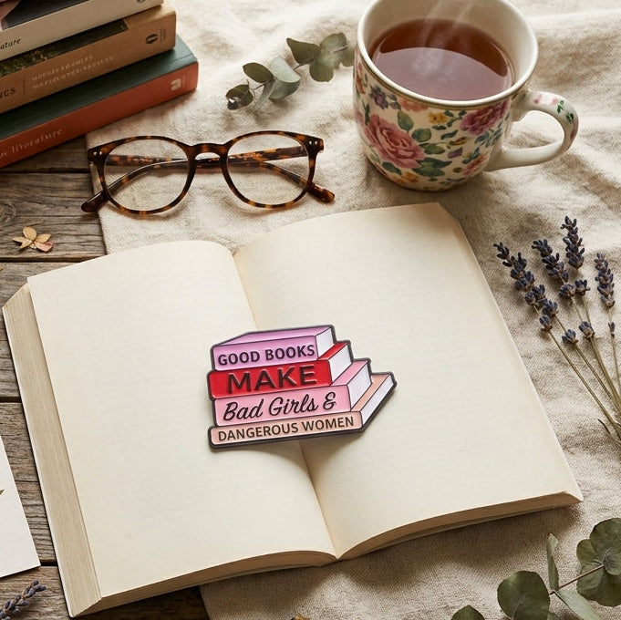 A styled flat-lay featuring a rectangular enamel pin with the text 'GOOD BOOKS MAKE Bad Girls & Dangerous Women' pinned to an open book. The scene is decorated with a canvas tote bag, a stack of books, a floral mug of tea, glasses, a notebook, and botanical stickers and sprigs on a rustic wooden table.