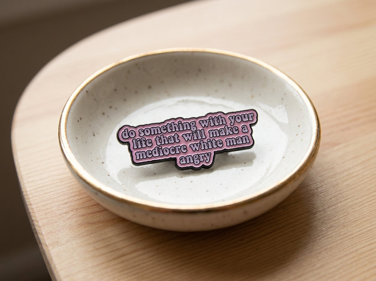 A pink enamel pin with a quote which reads 'do something with your life that makes a mediocre white man angry', resting inside a small speckled ceramic dish on a wooden surface.