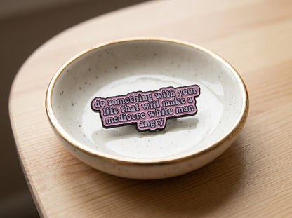 A pink enamel pin with a quote which reads 'do something with your life that makes a mediocre white man angry', resting inside a small speckled ceramic dish on a wooden surface.