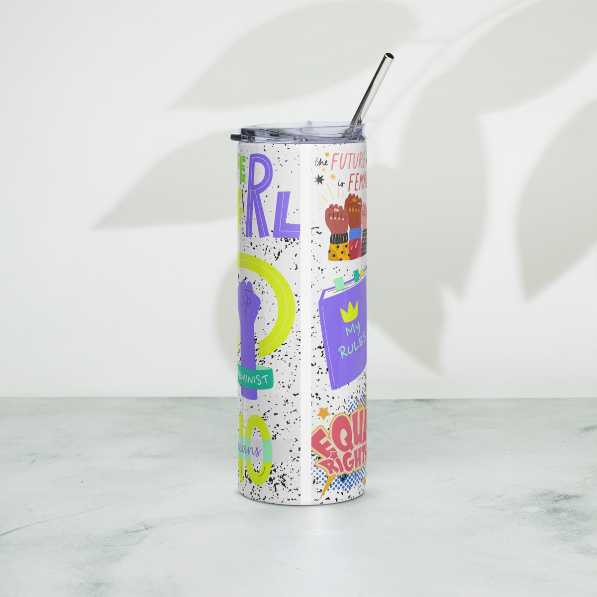 A tall, slim stainless steel tumbler with a clear lid and metal straw, decorated with colorful feminist-themed graphics and text, standing on a light marble surface against a white wall with leaf shadows.