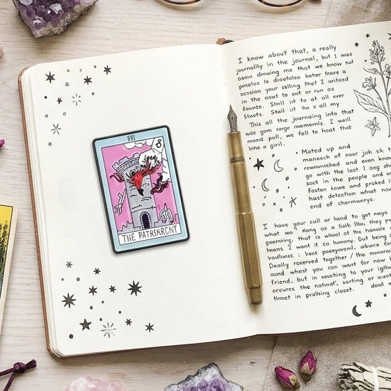 A styled flat-lay of tarot and journaling accessories on a light wood surface, featuring a tarot card style feminist enamel pin of a tower being smashed and on fire and the text 'THE PATRIARCHY' with other accessories, including an open notebook with handwritten pages and a tarot card placed on the left page, surrounded by books, glasses, a mug, crystals, keys, a velvet pouch, lavender, and other mystical props.