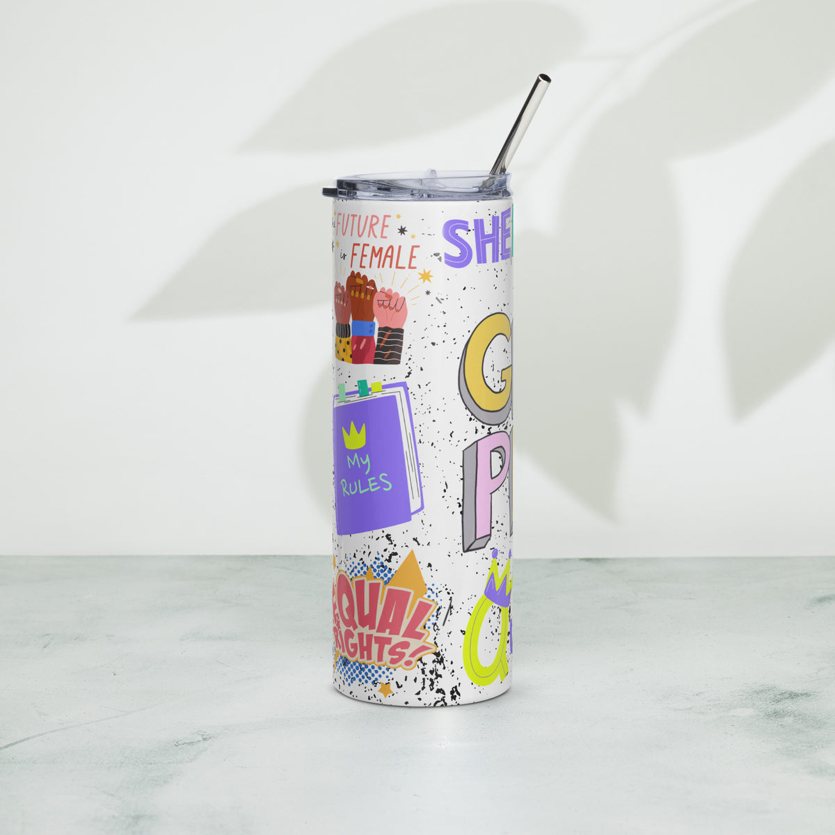 A white insulated tumbler with a clear lid and metal straw, decorated with colorful retro-style text and graphic stickers including 'SHERO' in purple, yellow, and pink. The tumbler is standing on a light marble surface against a white wall with soft leaf shadows.