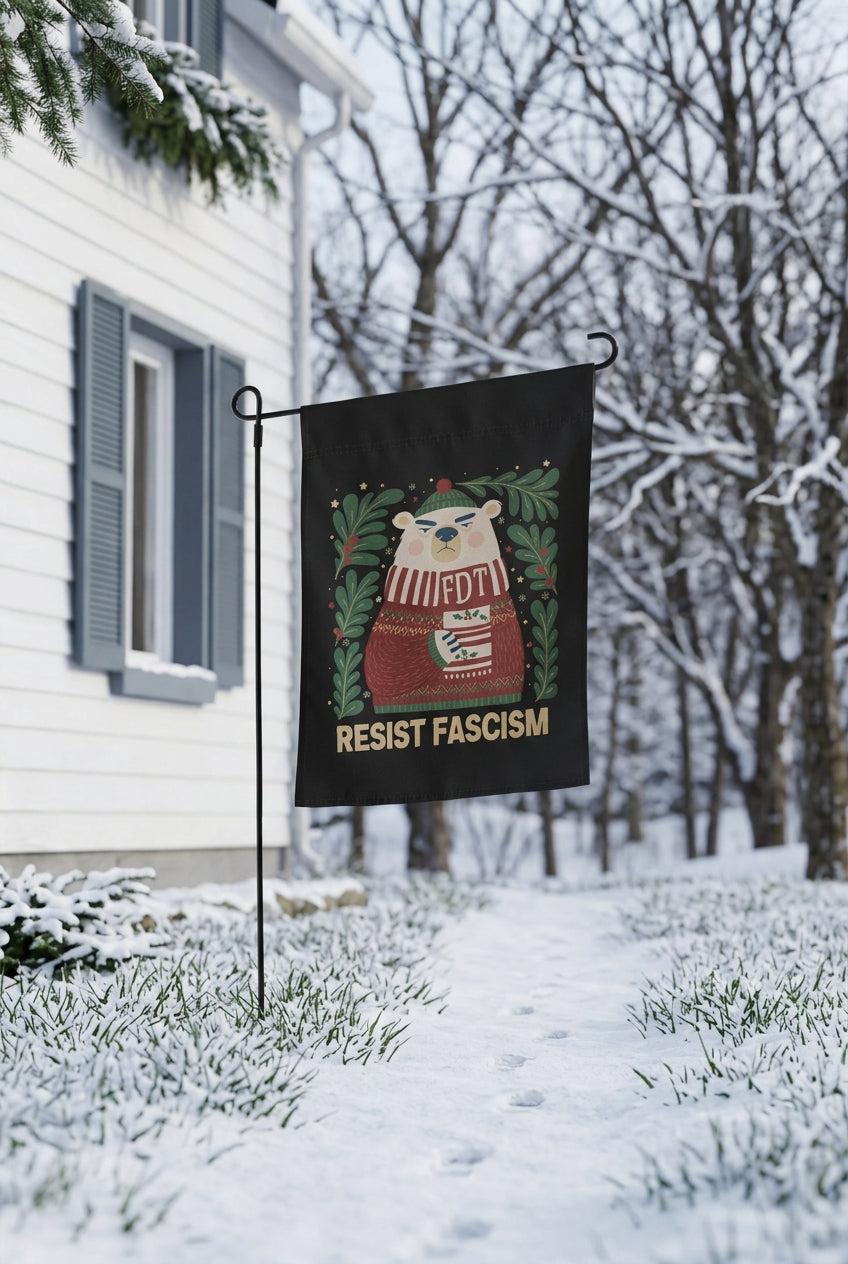 A black garden flag with a cartoon polar bear wearing a red sweater and holding a mug that says 'FDT', surrounded by green wreaths, with the text 'RESIST FASCISM' below. The flag is displayed on a metal stand in a snow-covered yard with a white house in the background.