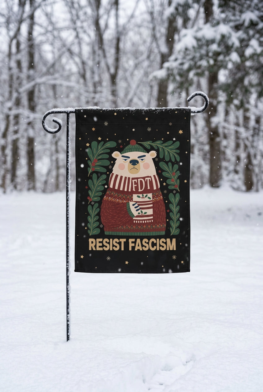 A black garden flag hanging on a black metal stake outdoors in the snow, with a cartoon bear wearing a sweater and the text 'FDT' and 'RESIST FASCISM' printed on it. The background shows a blurred park scene with trees and a path.