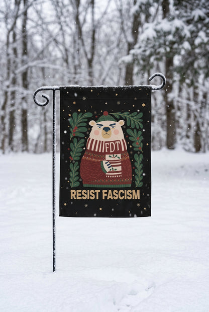 A black garden flag hanging on a black metal stake outdoors in the snow, with a cartoon bear wearing a sweater and the text 'FDT' and 'RESIST FASCISM' printed on it. The background shows a blurred park scene with trees and a path.