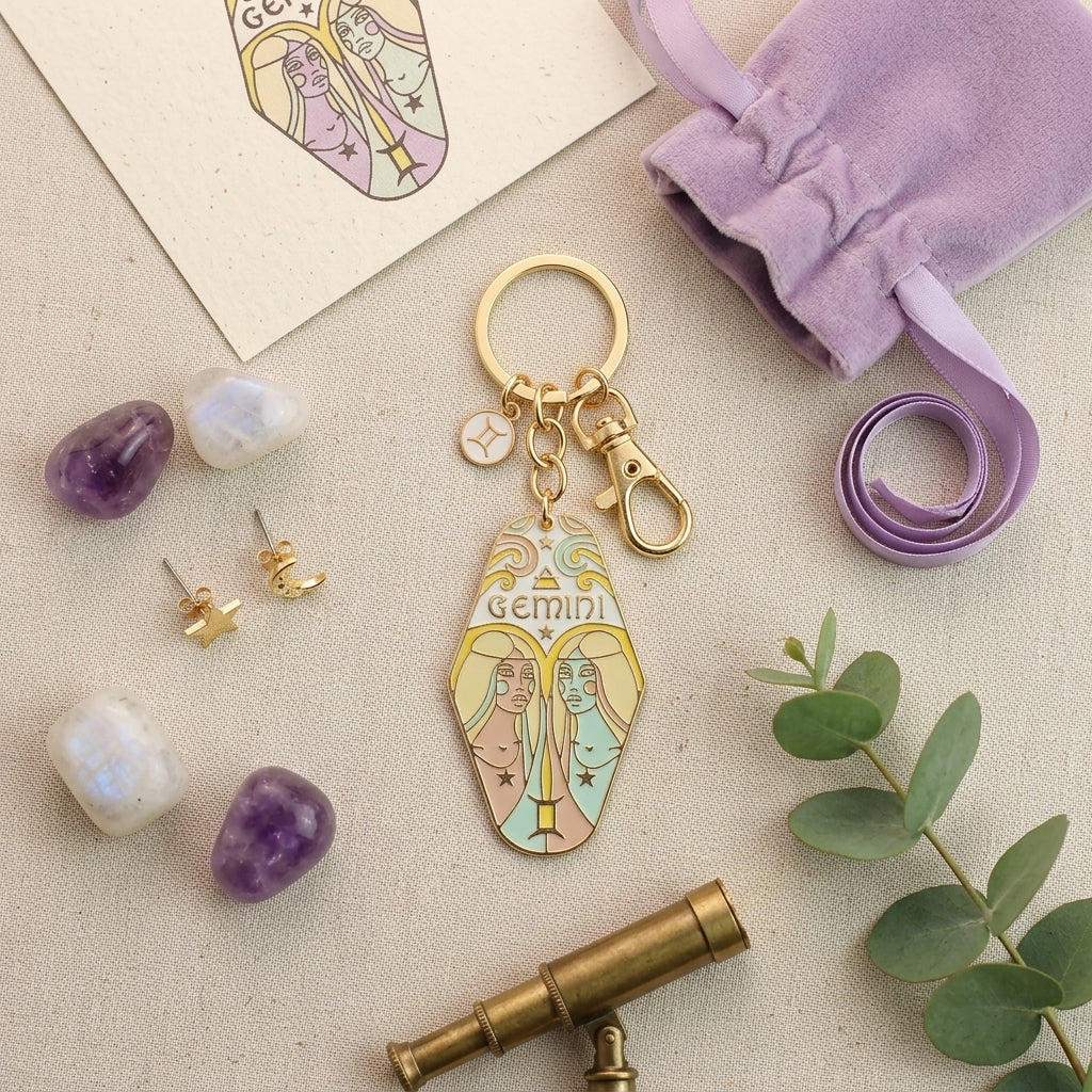 A gold-tone Gemini zodiac keychain charm with pastel-colored illustrations of twin Gemini figures, styled in a flat-lay arrangement with a lavender drawstring bag, crystals, earrings, a telescope, and green leaves on a beige fabric background.