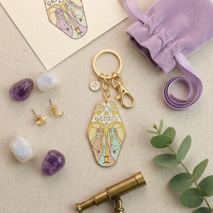 A gold-tone Gemini zodiac keychain charm with pastel-colored illustrations of twin Gemini figures, styled in a flat-lay arrangement with a lavender drawstring bag, crystals, earrings, a telescope, and green leaves on a beige fabric background.