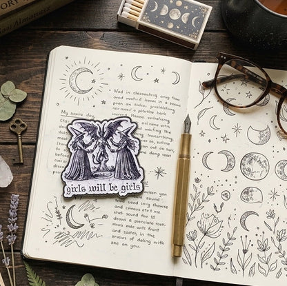 An open journal with moon and star illustrations lies on a wooden table, featuring a central enamel pin of two witches with the text 'girls will be girls'. The scene is styled with a tarot card, amethyst crystals, a matchbox, glasses, a cup of tea, a pen, and other mystical accessories.