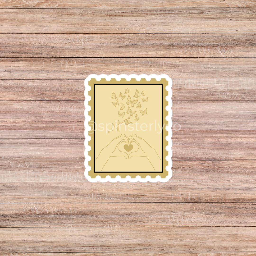 A rectangular decorative print or sticker with a scalloped white border, featuring a pale yellow background, a black inner border, and a gold line drawing of hands forming a heart with butterflies above it. The item is centered on a brown wood plank background.