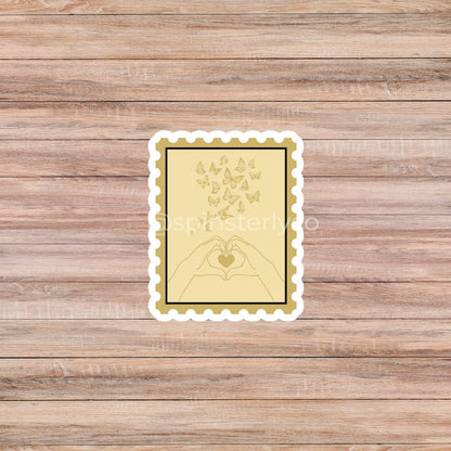 A rectangular decorative print or sticker with a scalloped white border, featuring a pale yellow background, a black inner border, and a gold line drawing of hands forming a heart with butterflies above it. The item is centered on a brown wood plank background.