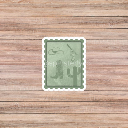 A rectangular postage-stamp-style sticker with a white scalloped border, shown centered on a light brown wood plank background. The sticker has a muted sage green face with western-themed line drawings including a cowboy hat, boots, a revolver, and a cactus.