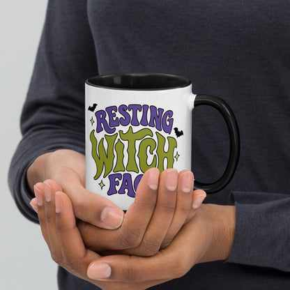 A person holding a white ceramic mug with a black handle and black interior. The mug features the text 'RESTING WITCH FACE' in purple and green lettering with small black bat silhouettes, shown against a neutral gray background.