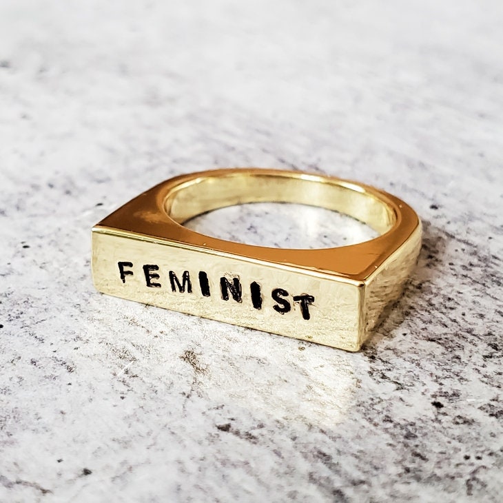 Gold ring with 'FEMINIST' engraving on a marble surface