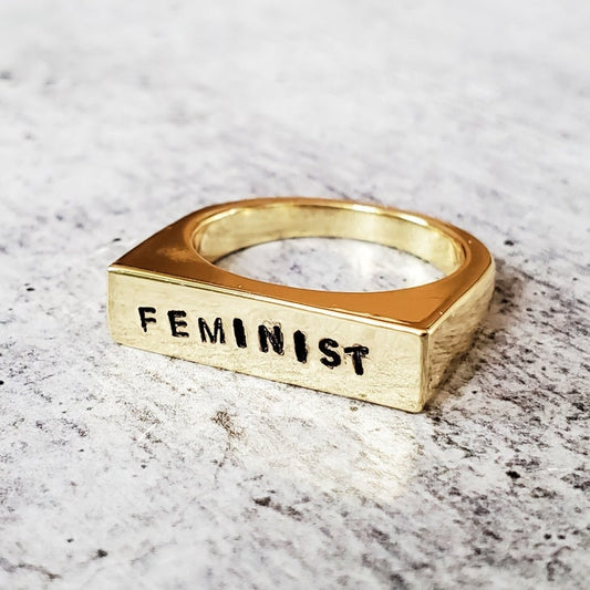 Gold ring with 'FEMINIST' engraving on a marble surface