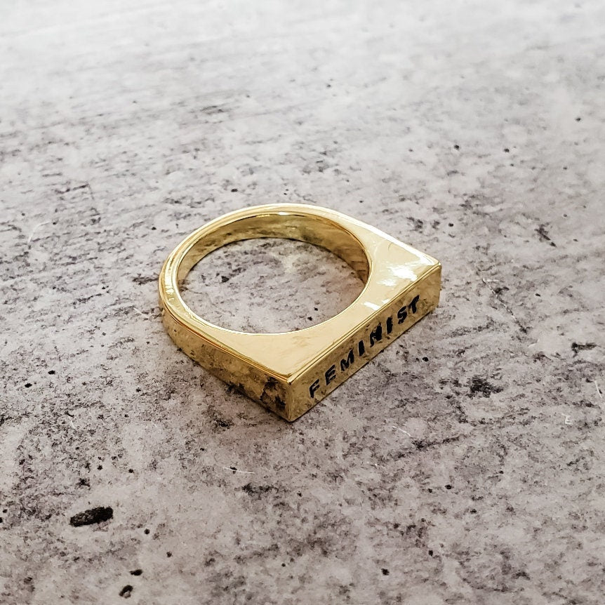 Gold ring with 'FEMINIST' engraving on a textured surface