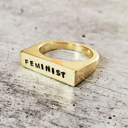 Gold ring with 'FEMINIST' engraving on a textured surface